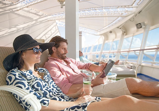 Woman wearing hat and sunglasses, holding glass of cucumber water, next to man wearing shorts and button down shirt holding iPad in lounge chairs in the Sanctuary